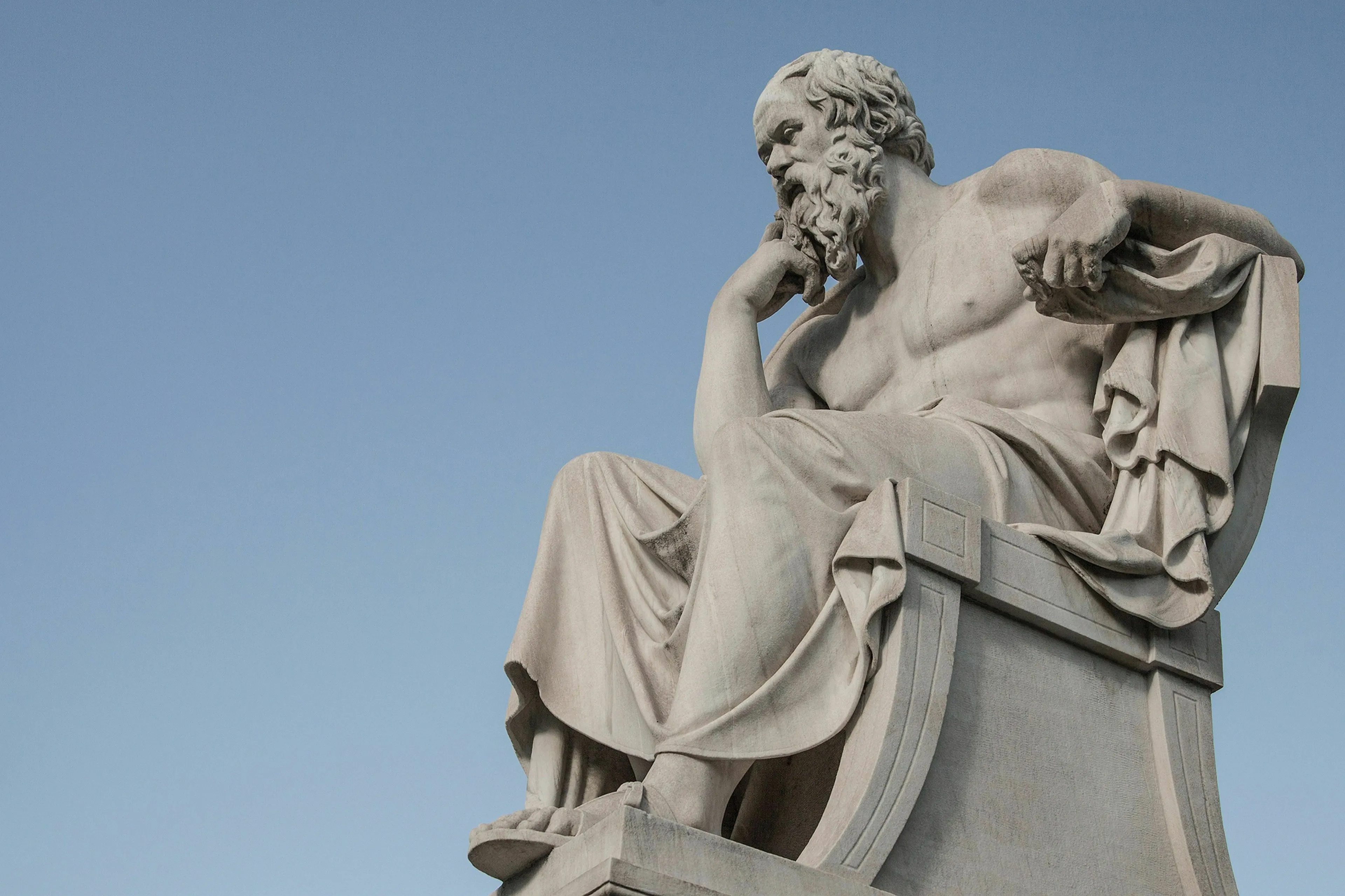 A stone statue of a seated philosopher, resembling Socrates, deep in thought, against a clear blue sky. Test your general knowledge.