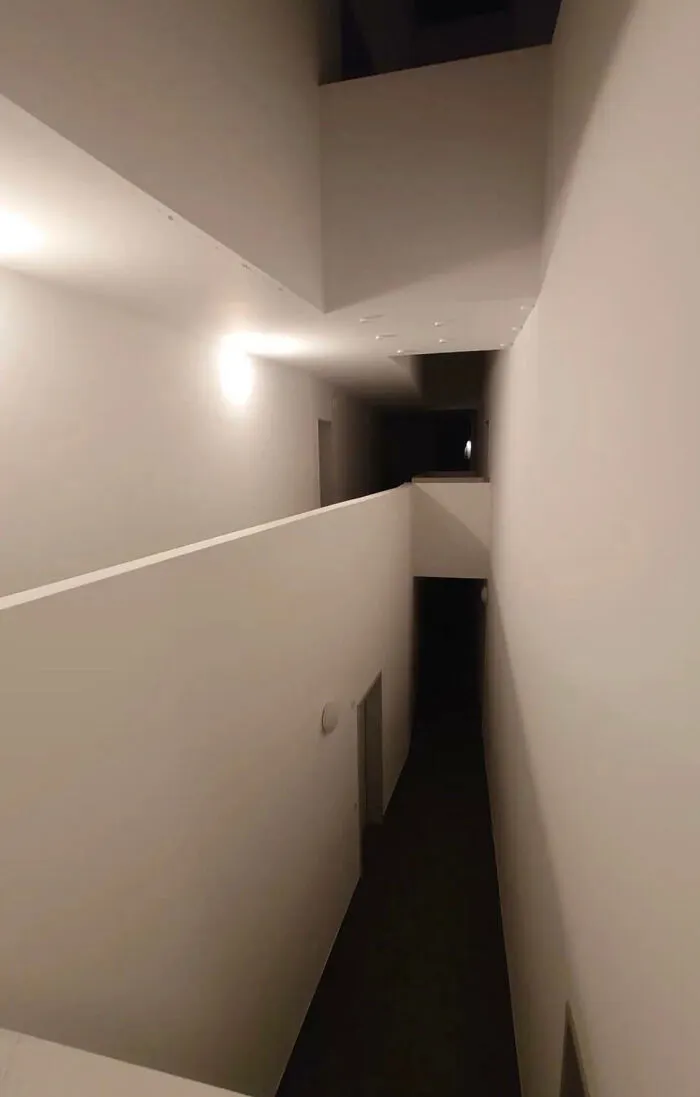 A narrow, stark white hallway with a dark, descending path, embodying a deeply unsettling liminal space.