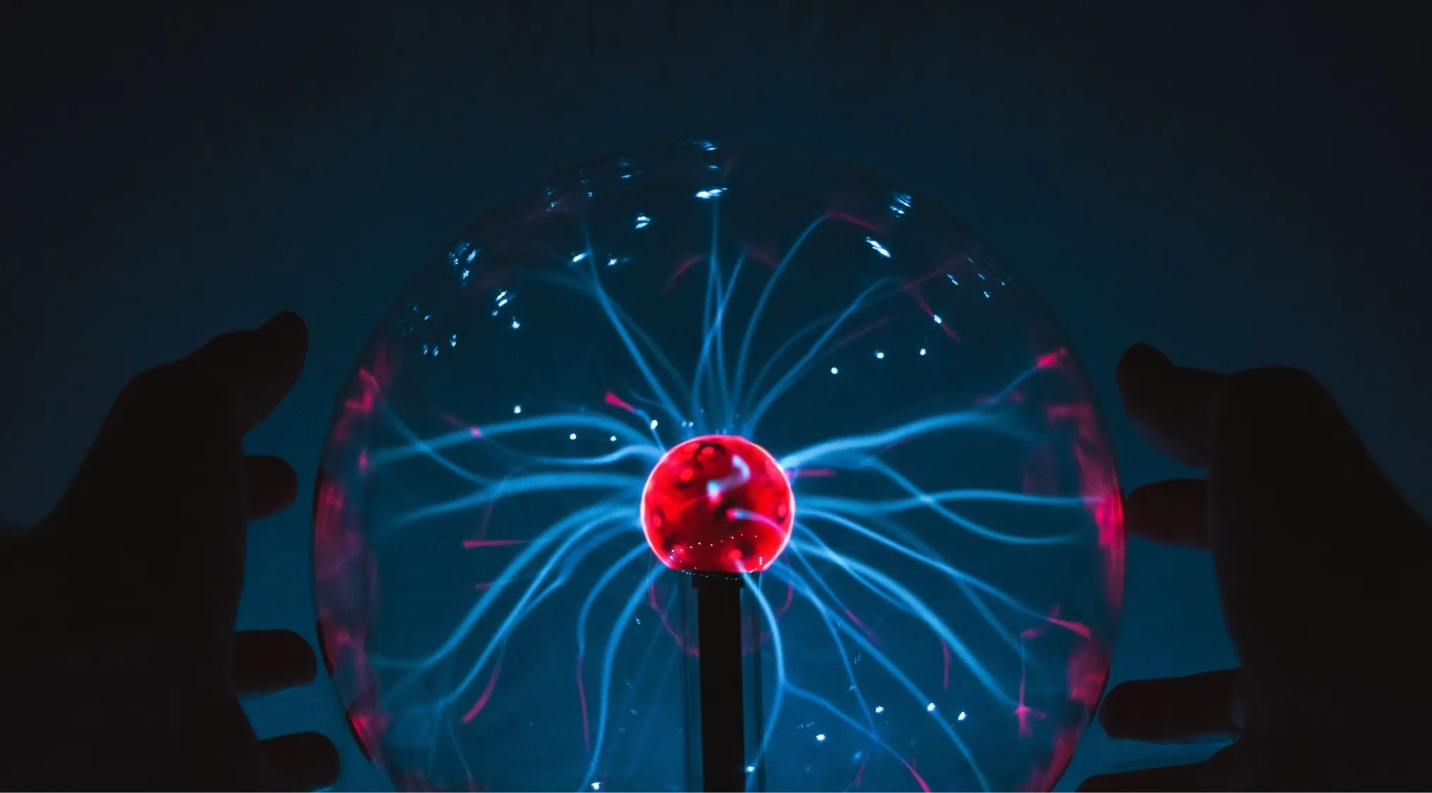 Hands touching a plasma globe, with blue and red electrical streams emanating from the center, illustrating science and energy.