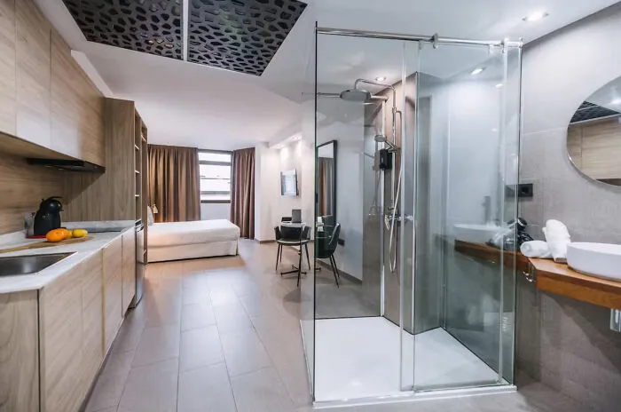 An unhinged real estate listing featuring an open-concept studio apartment with a bed, kitchen, and glass shower.