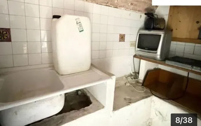 A small, grimy utility room with an old sink, a large white plastic container, and an outdated TV, part of unhinged real estate listings.