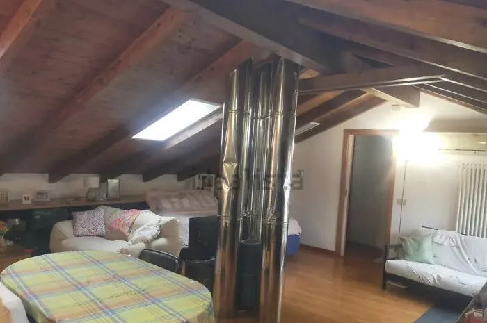 Unhinged real estate listing: an attic living space with exposed wooden beams and a large, shiny metal chimney pipe dominating the center.