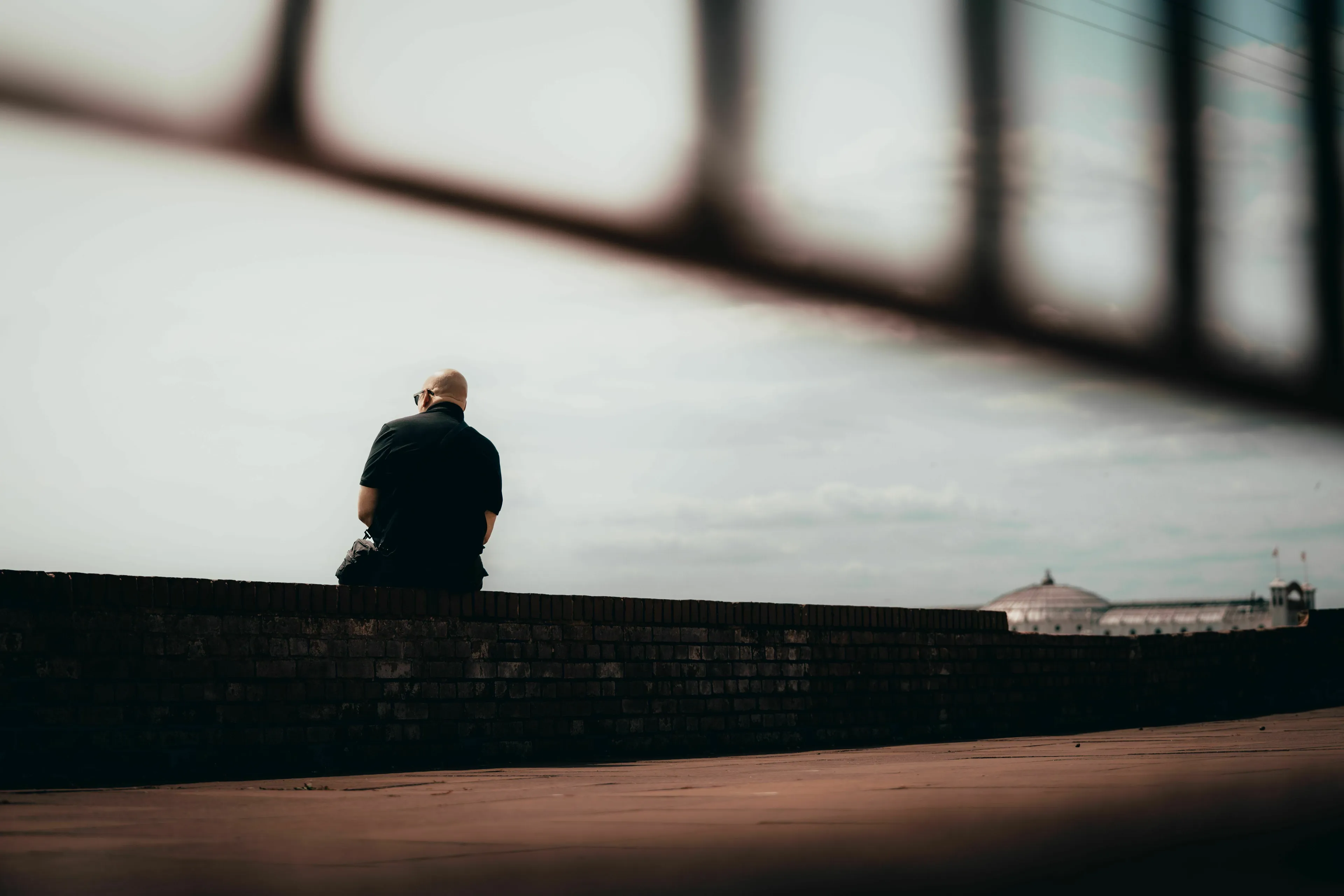 A lone man sits on a brick wall, back to the camera, looking out at the distant horizon, contemplating loneliness.