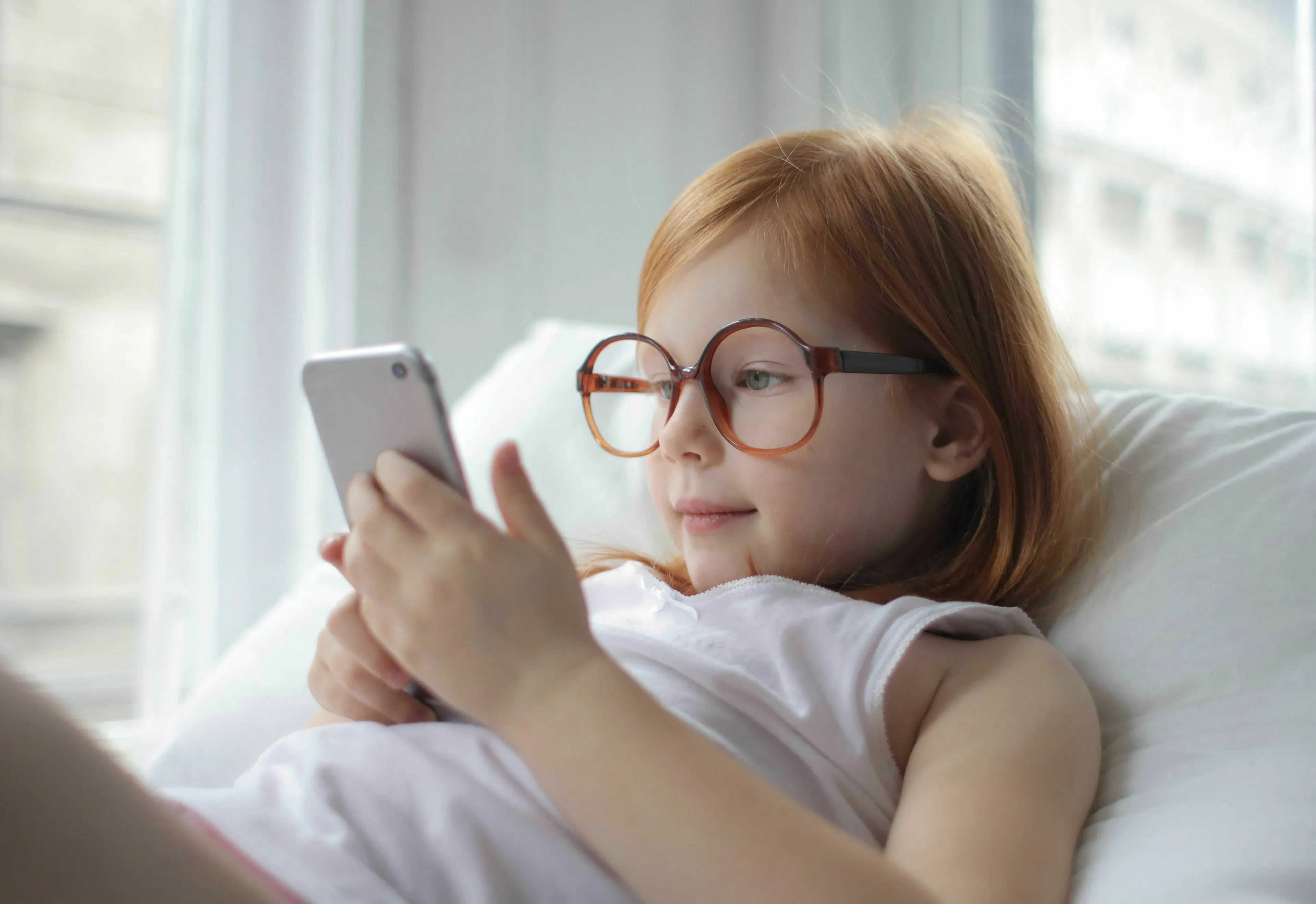 A young girl with glasses lying down and smiling while holding a smartphone, ideal for handling loneliness.