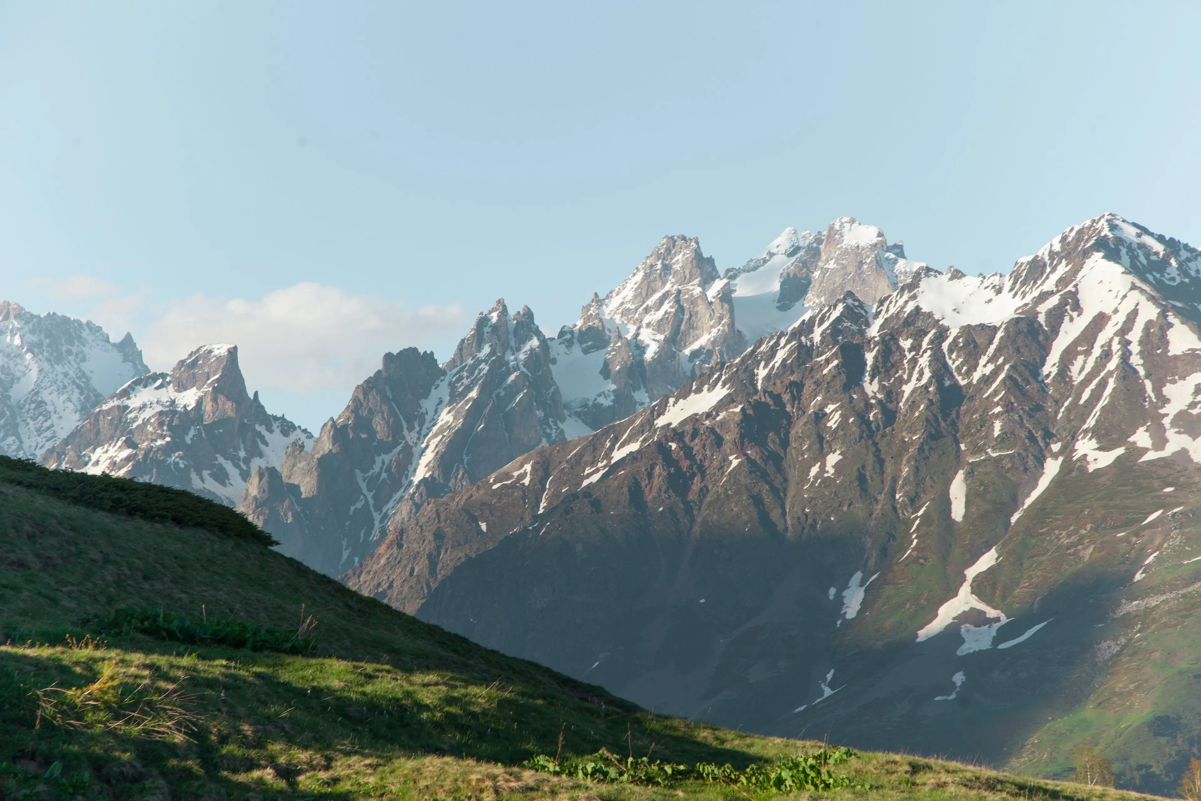 Towering mountains, snow-capped peaks against a clear sky, with green hills in the foreground.