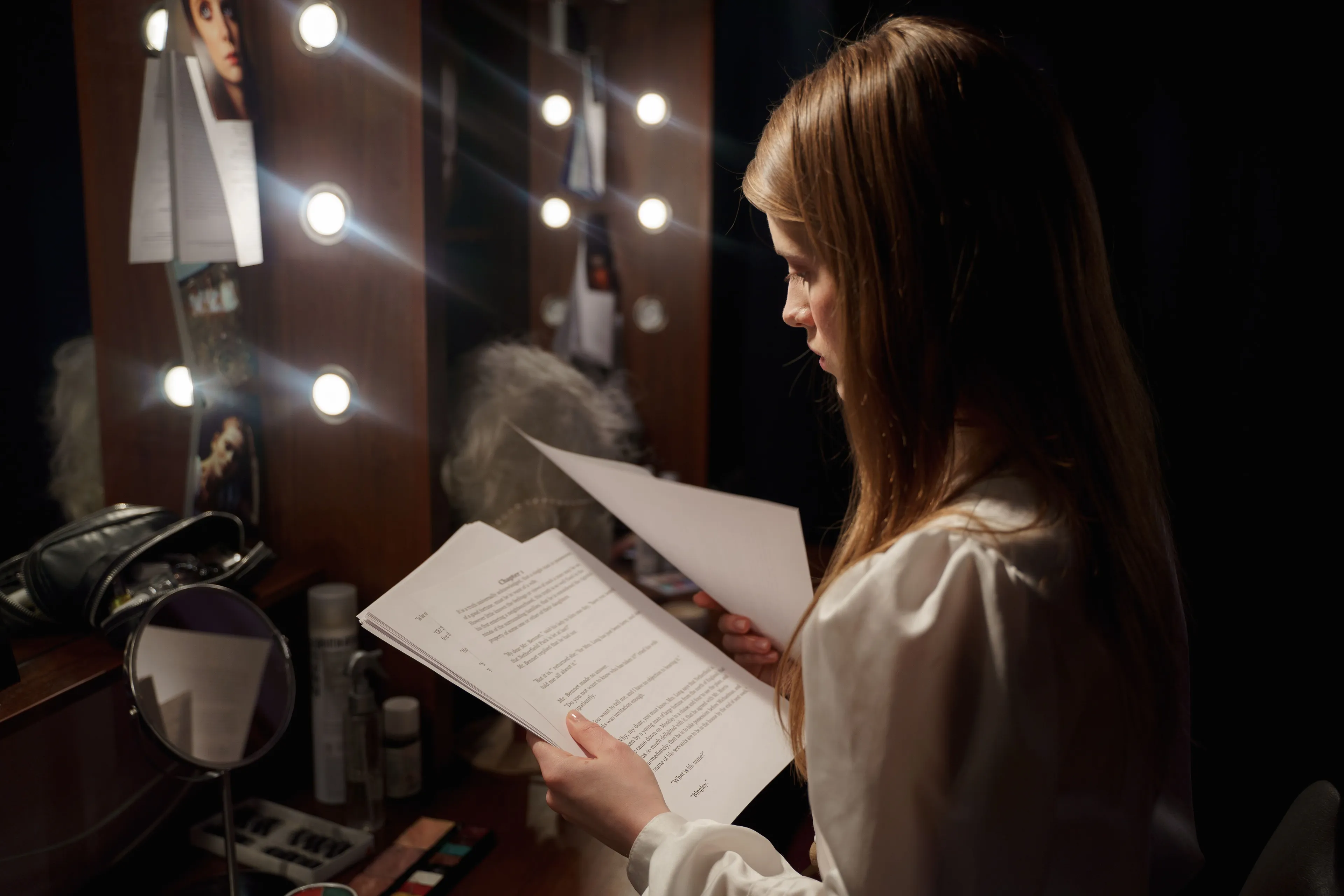 A young woman, a playwright, reviewing scripts in a dimly lit dressing room with lit vanity bulbs.
