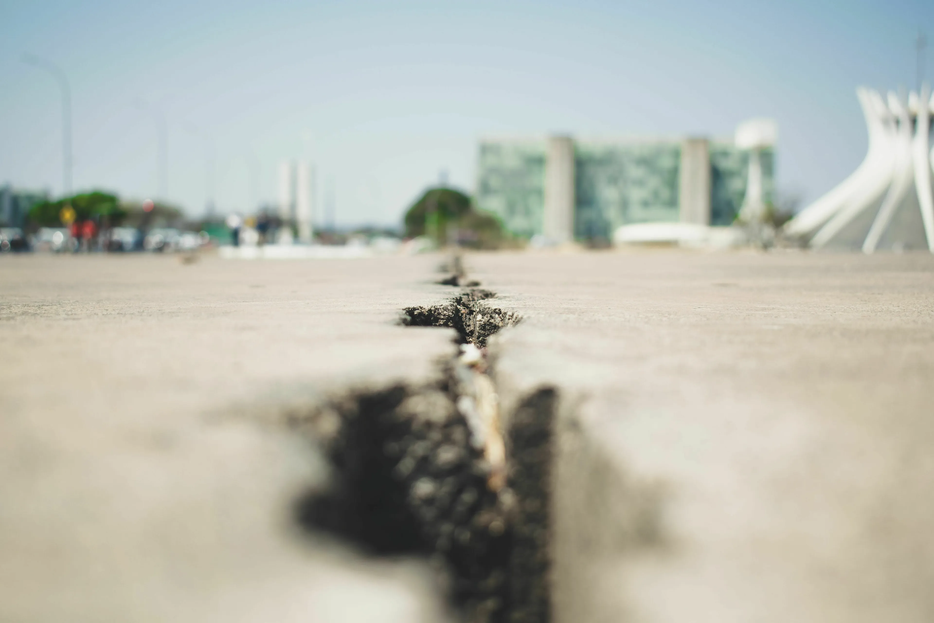A close-up, low-angle shot of a crack in the ground, with a blurred background of a modern building, hinting at earthquakes.
