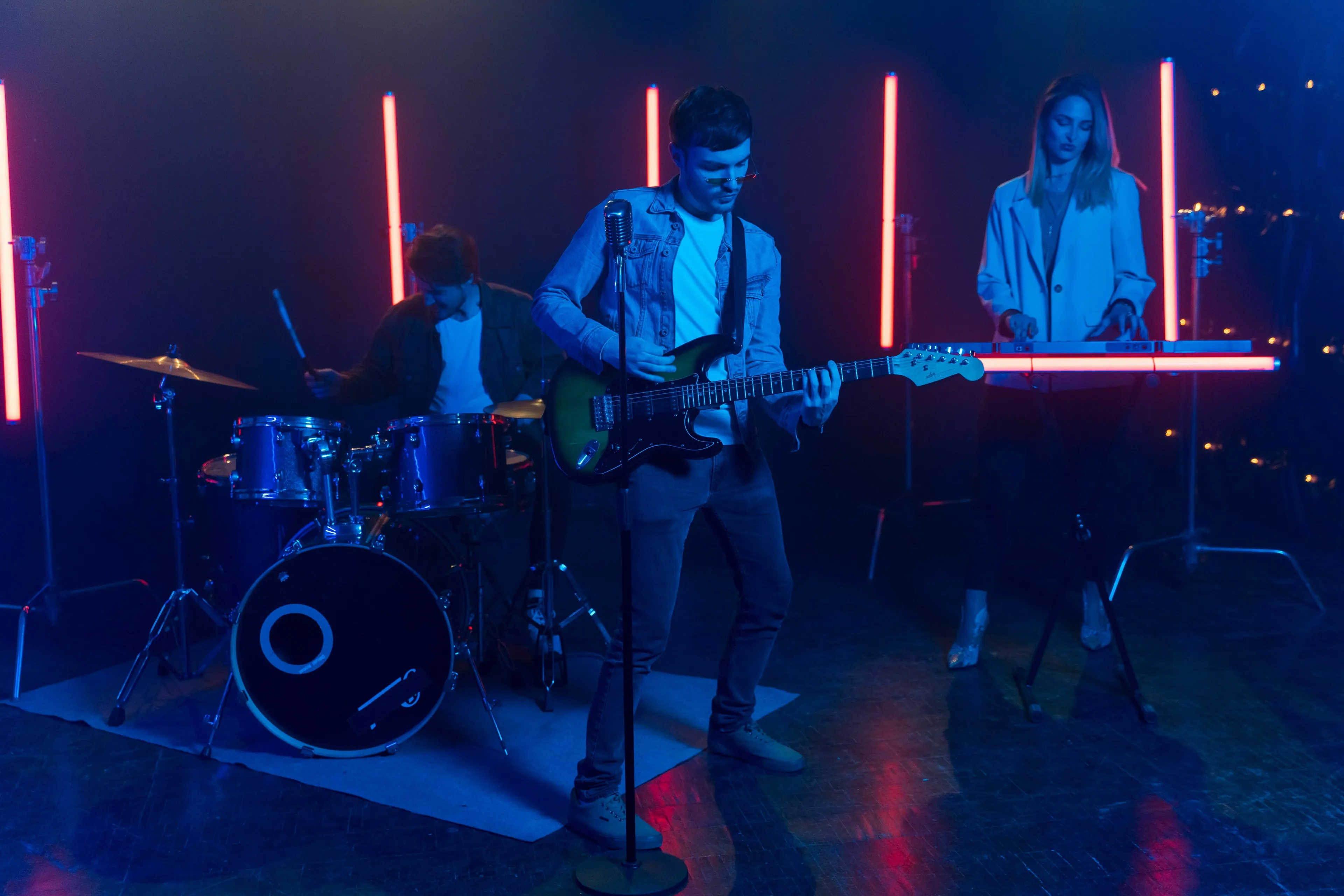 A band performing on stage with red neon lights in the background. A guitarist is at the center, a drummer to the left, and a keyboardist to the right, playing live music.