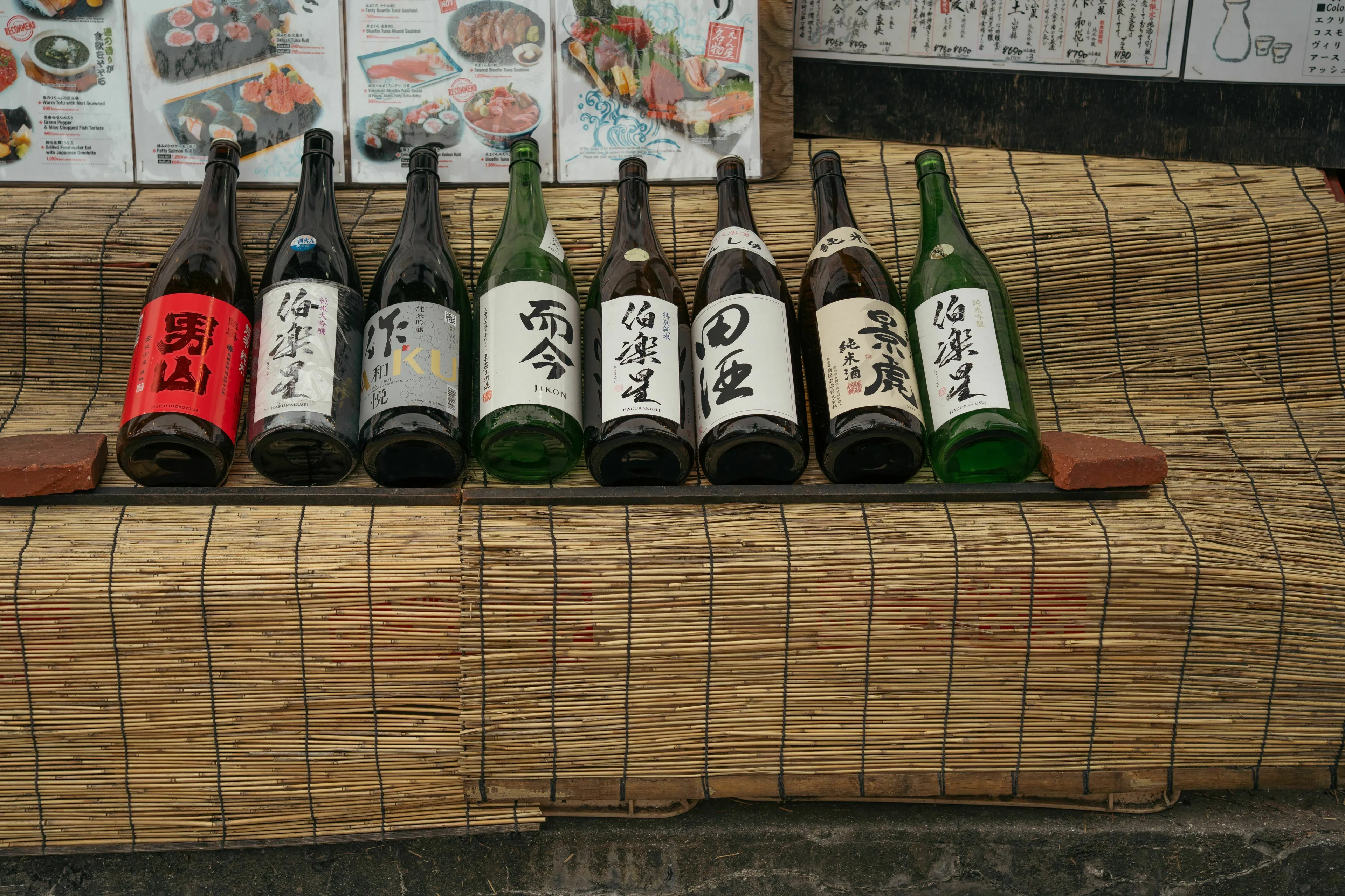 Several bottles of sake lined up on a straw mat in front of a menu. The bottles are mostly dark, with white labels, showcasing different sake varieties.