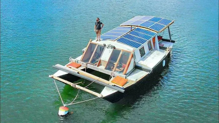 A man stands on the roof of a wacky houseboat equipped with solar panels, floating on calm blue water.