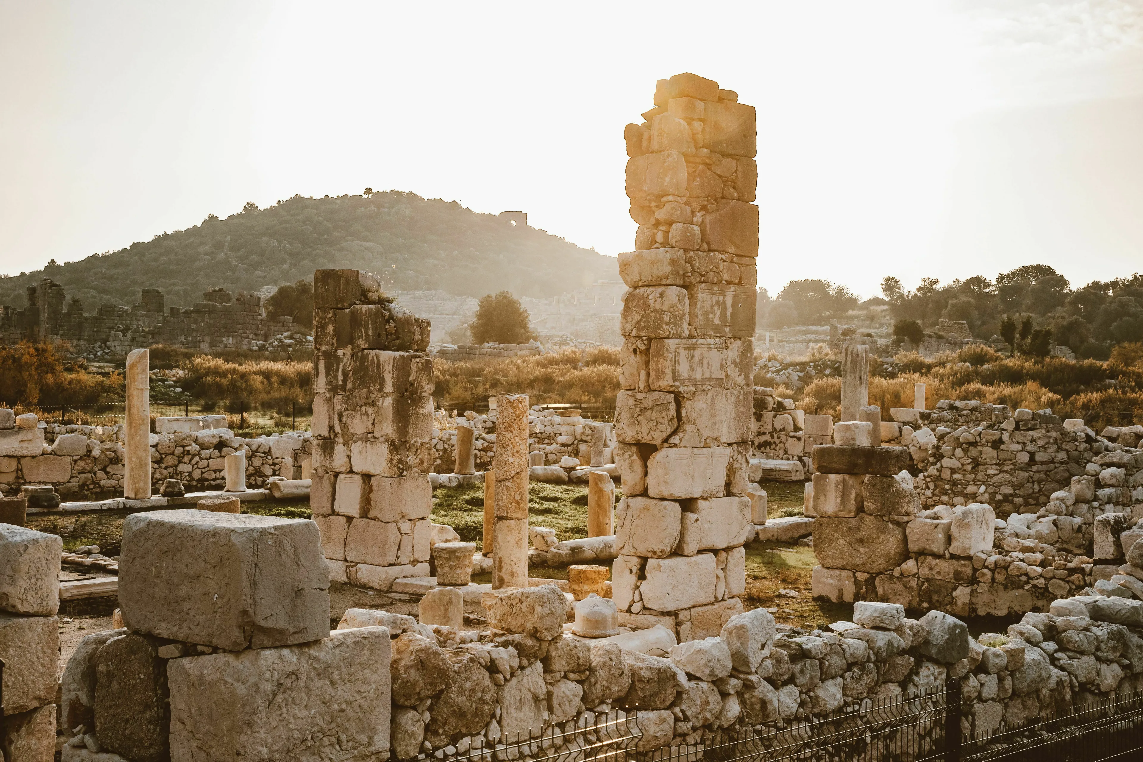 Ancient civilization ruins in a field with a hill in the background, showing stone columns and foundations at sunset.