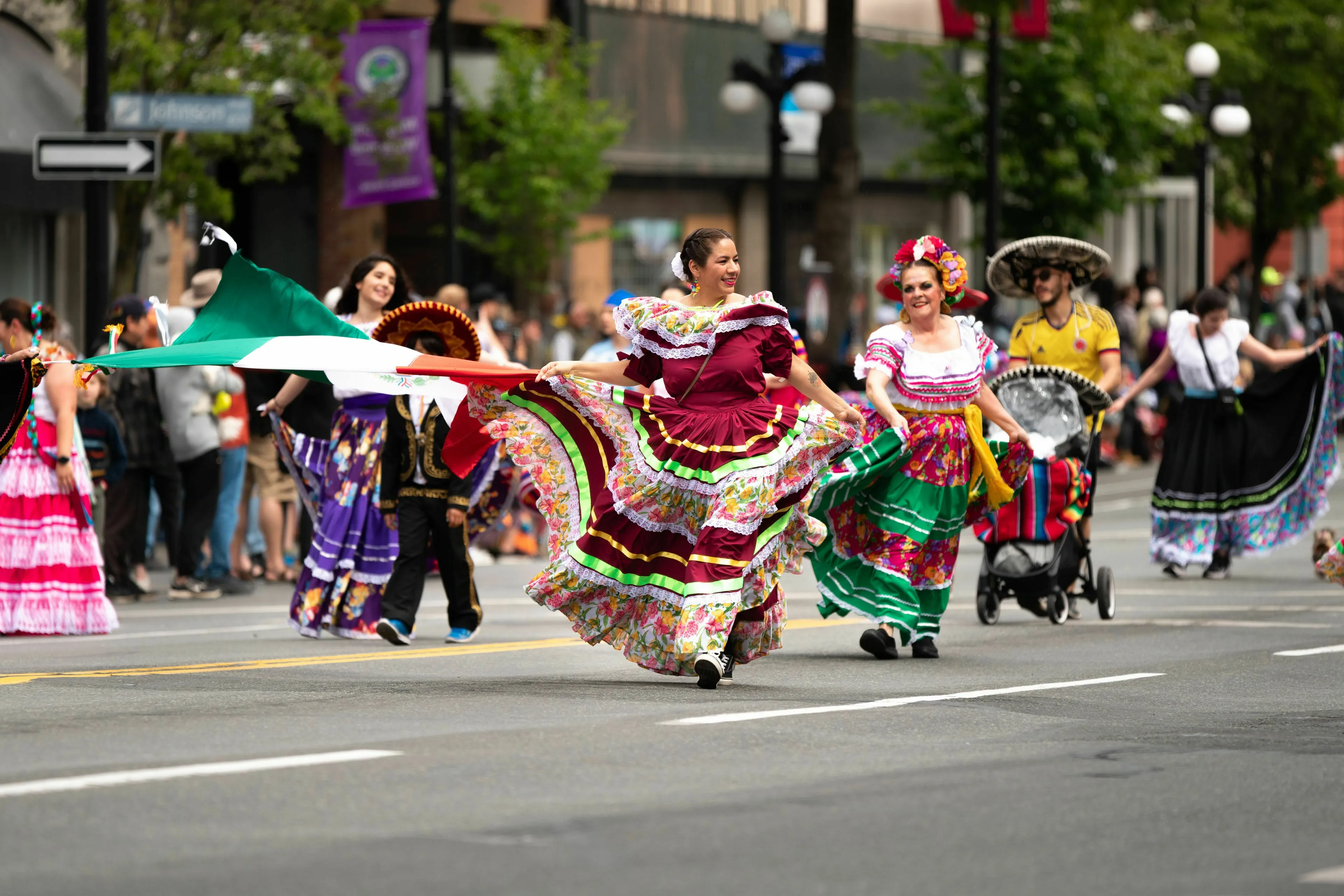 People in traditional folk dresses carrying a Mexican flag during a festival day parade on a city street.
