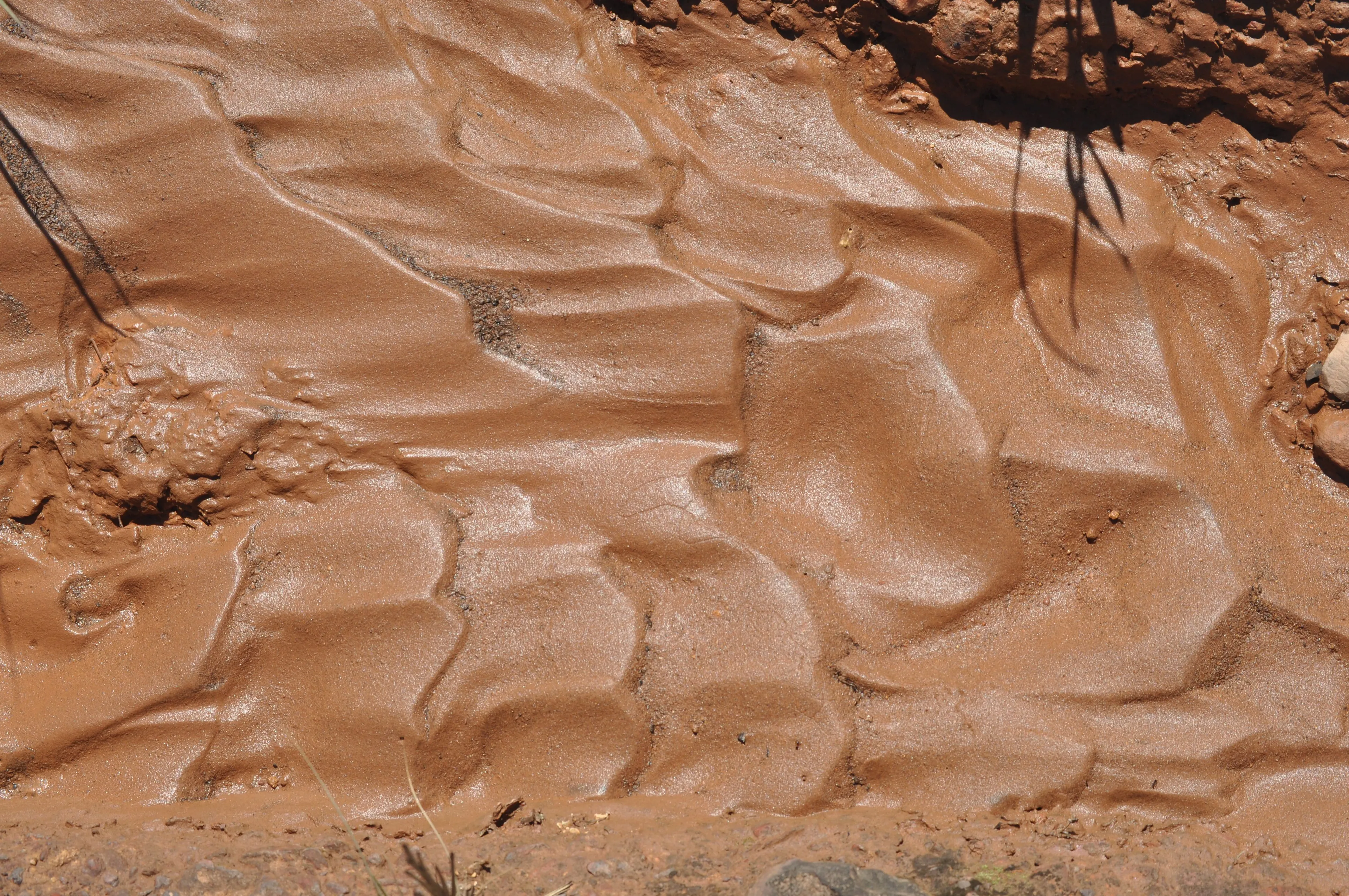 Close-up of wet, reddish-brown clay showing intricate patterns formed by water flow. The clay material is smooth and shiny.