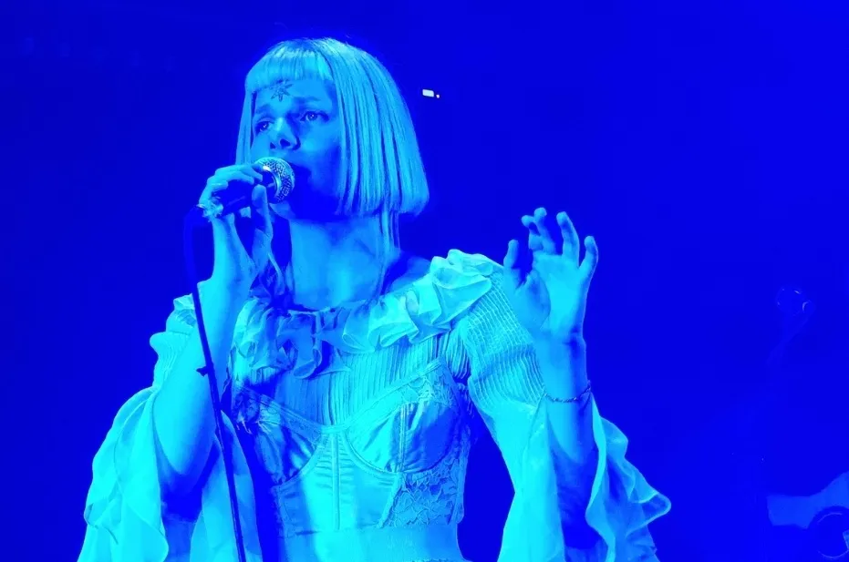 Icelandic musician Aurora performing on stage, bathed in blue light, holding a microphone, with a star on her forehead.