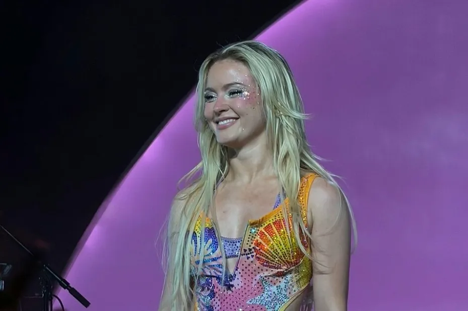 A smiling Zara Larsson with blonde hair and glitter makeup, wearing a colorful, bedazzled top on stage. Icelandic musician.