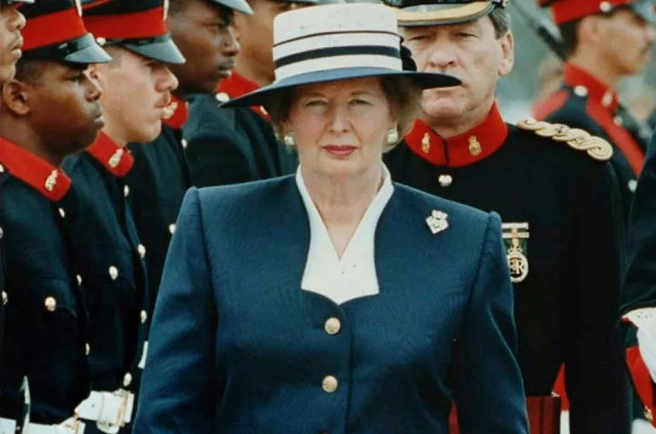 Margaret Thatcher, the UK's first female PM, in a white and blue hat and suit, standing among military officers.