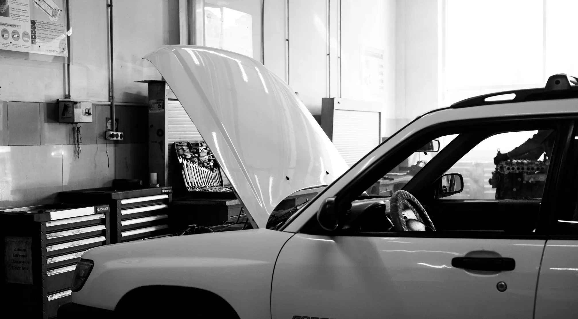 Black and white shot of a car with an open American hood in a garage. Tools and a car engine are visible.