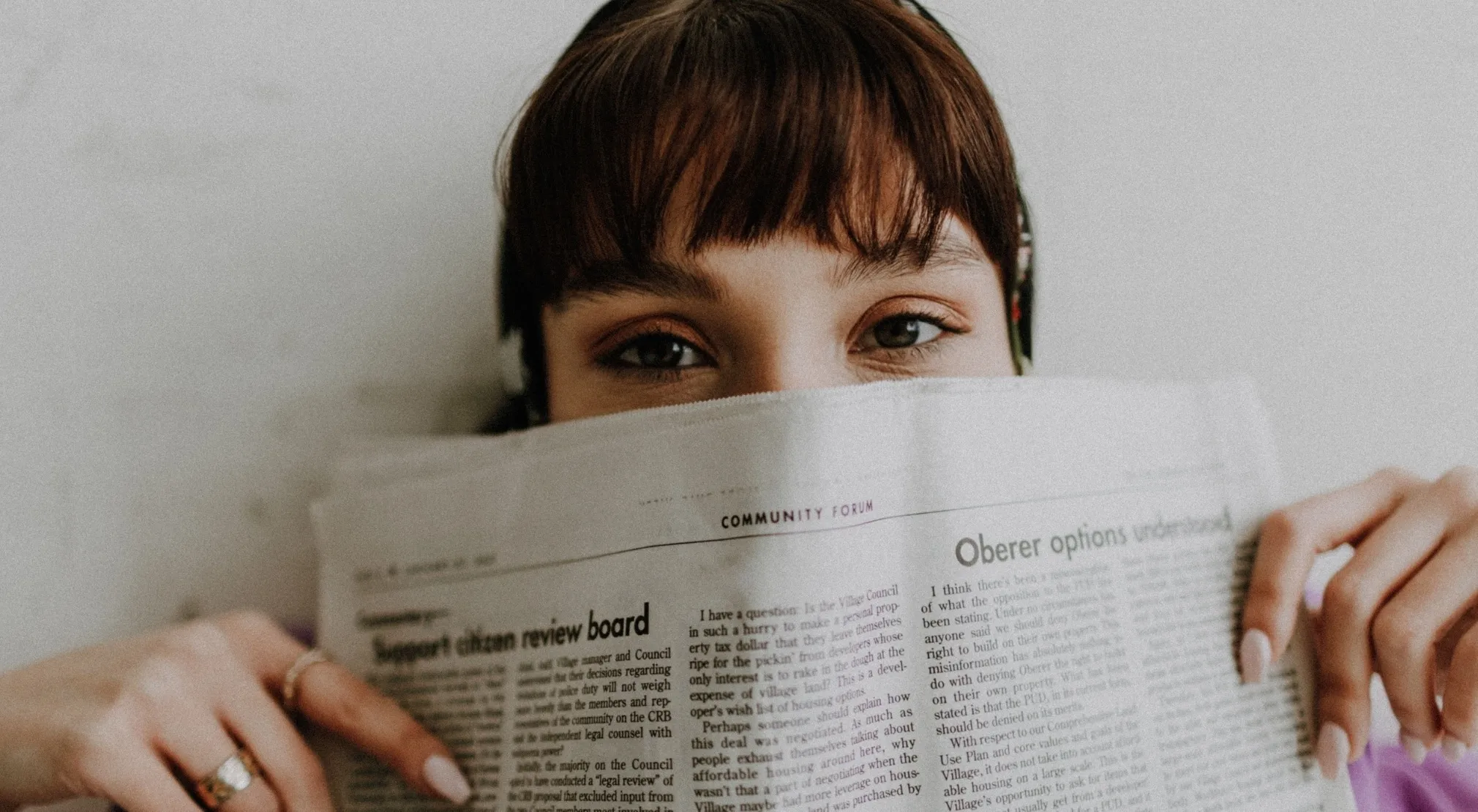 A woman with a fringe (American bangs hairstyle) hides behind a newspaper, with only her eyes and forehead visible.