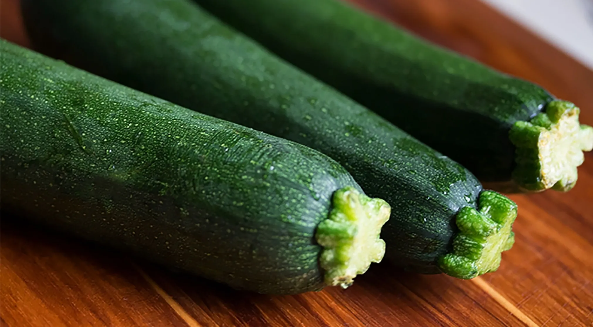 Three fresh green zucchini on a wooden cutting board. A Brit would call these courgettes, American vs Brit Words.
