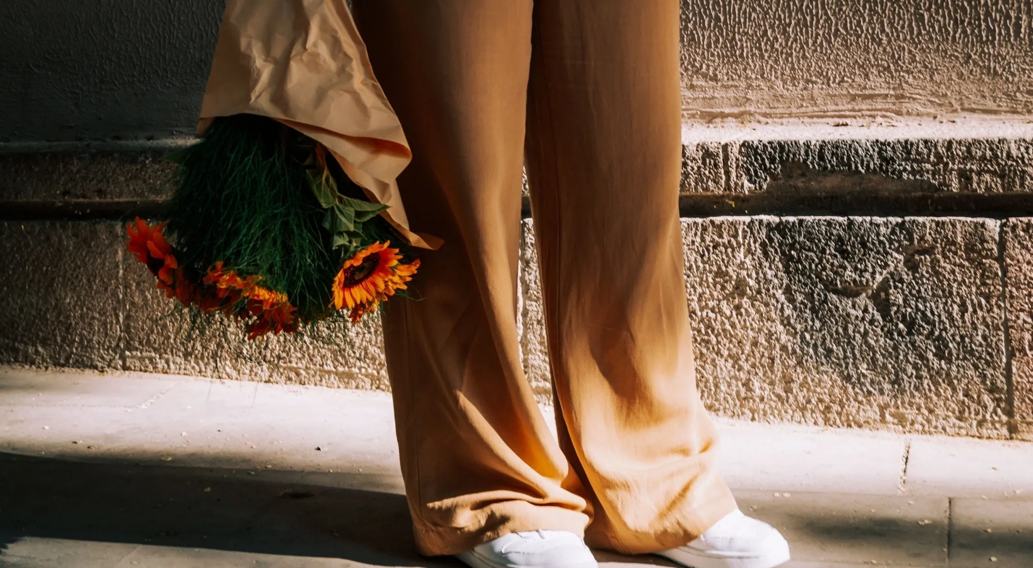 A person wearing brown wide-leg pants and white sneakers, holding a bouquet of orange flowers against a textured wall.