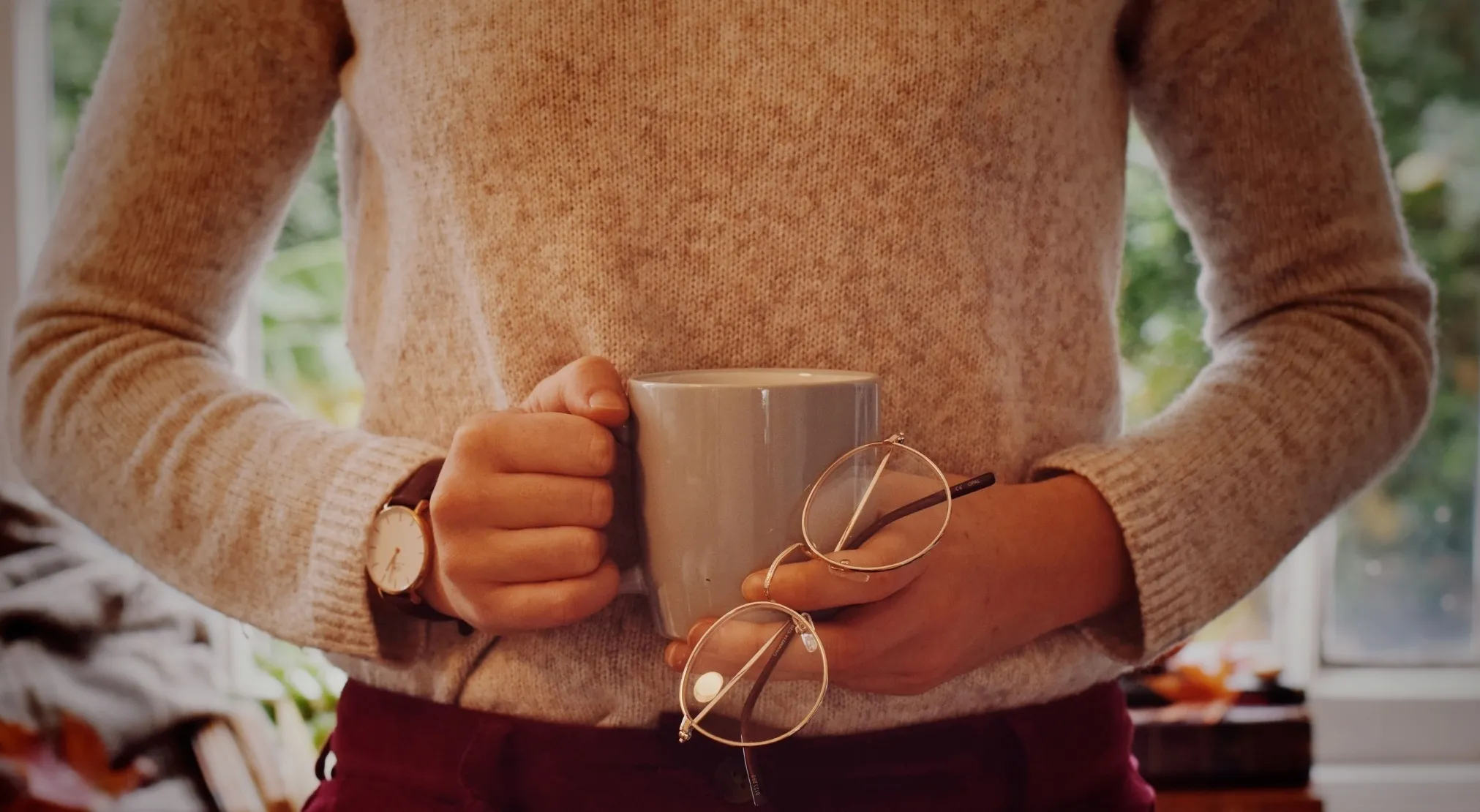 Close-up of a person in a beige American sweater, holding a mug and round glasses, in a cozy setting.