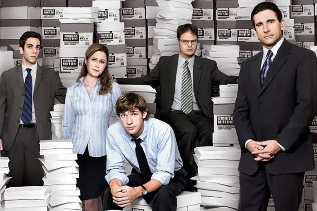 The cast of The Office posing in an office setting with stacks of paper in the background.