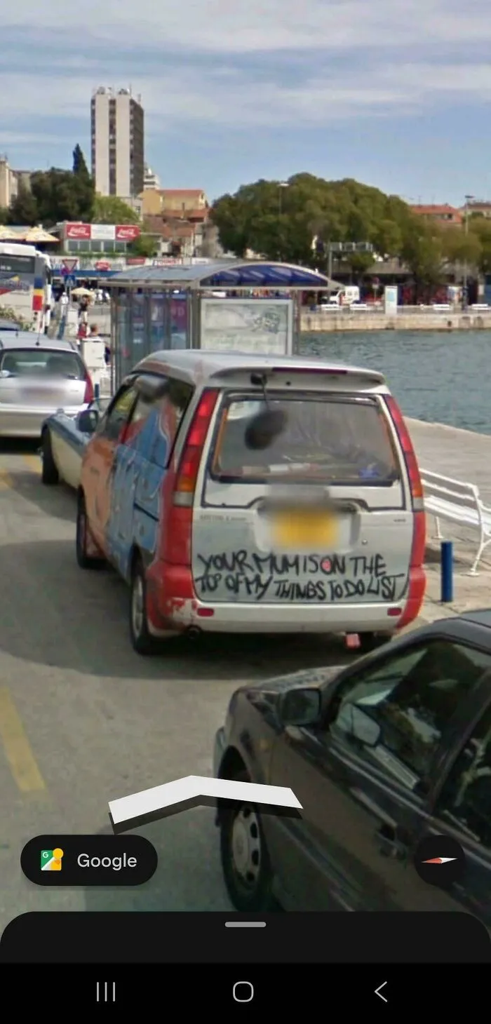 A car with graffiti on its back window parked by the water, seen in a funny Google Street View photo.