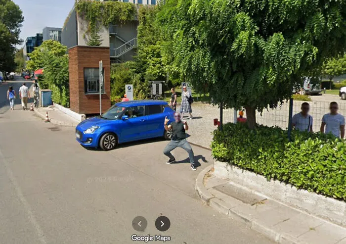 A man in a mask making a funny pose next to a blue car, captured in a Google Street View photo.