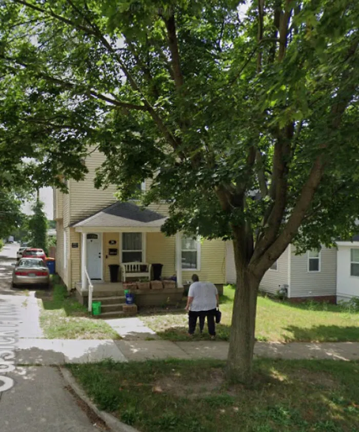 A person walks past a house, captured in a funny Google Street View photo, under a large tree.