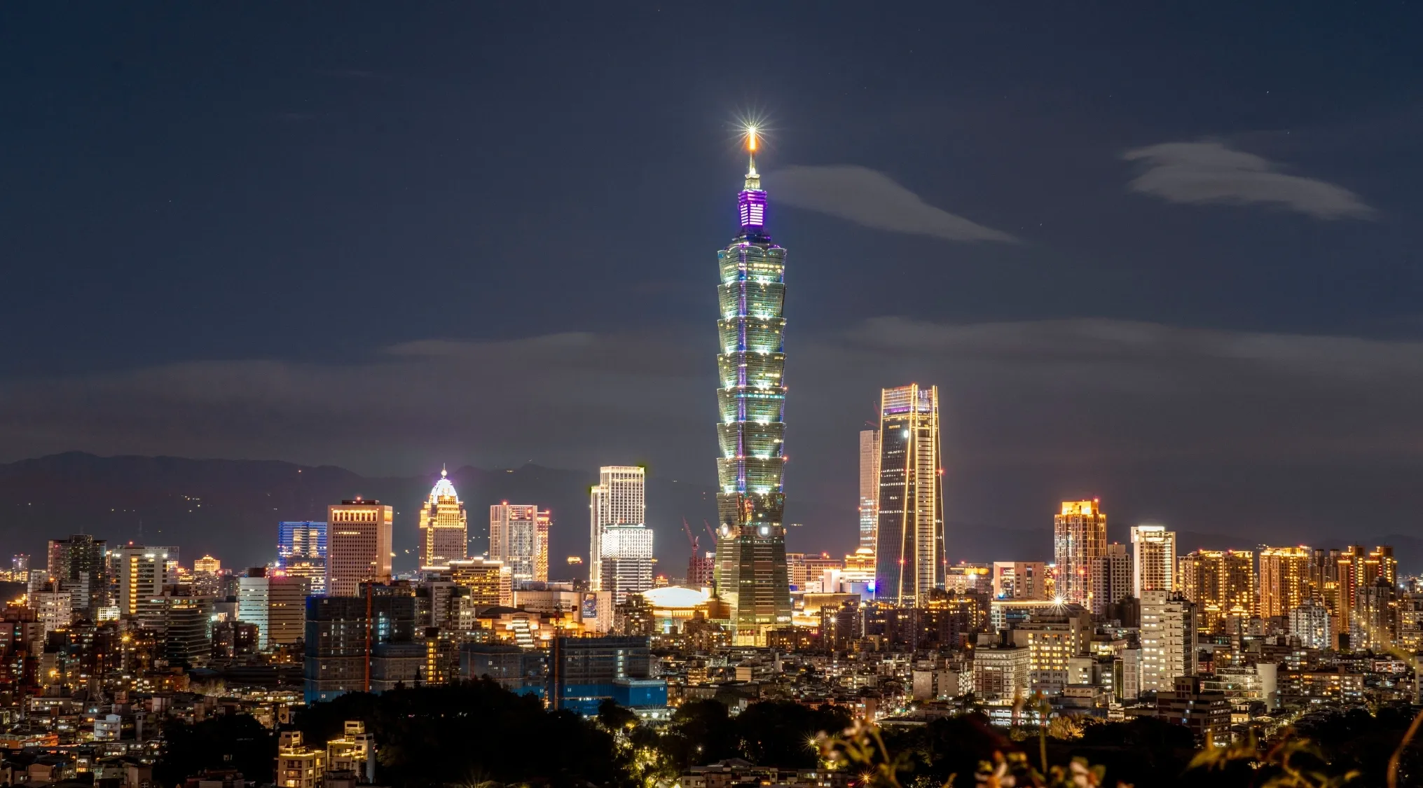 Taipei 101, a bamboo-like building, glowing with lights against a dark night sky, surrounded by a city skyline. Guess the country.