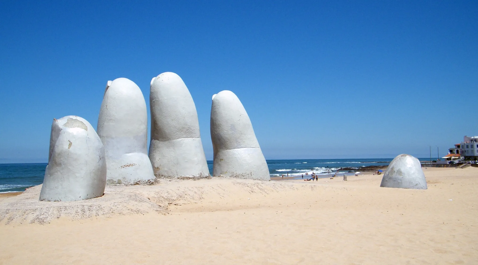 Guess the Country: La Mano, an iconic hand sculpture, emerging from a sandy beach with the ocean and blue sky in the background.