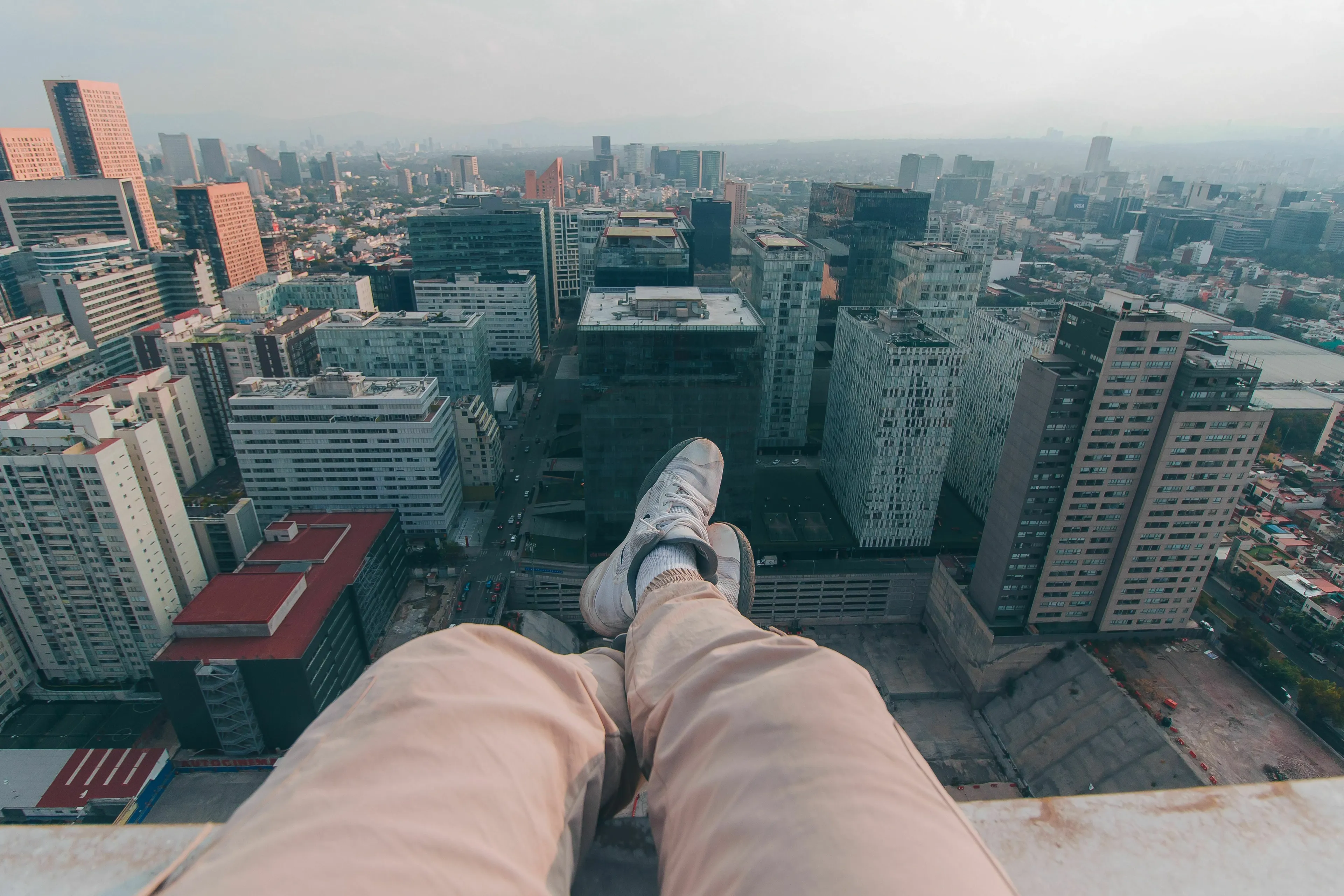 A person's legs dangling over the edge of a tall building, overlooking a city, representing heights.