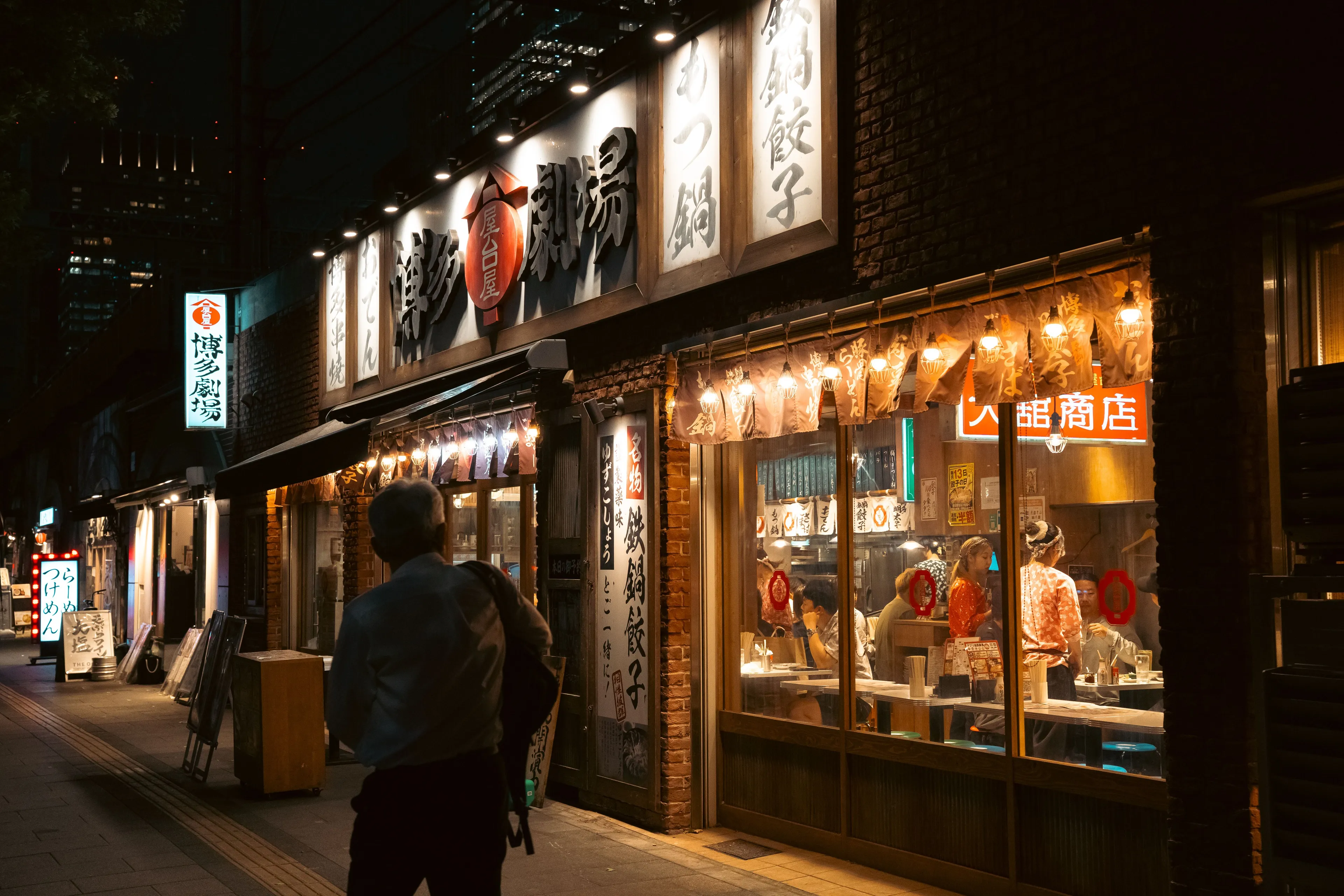 A night view of a brightly lit street with a new restaurant, as a man walks past.