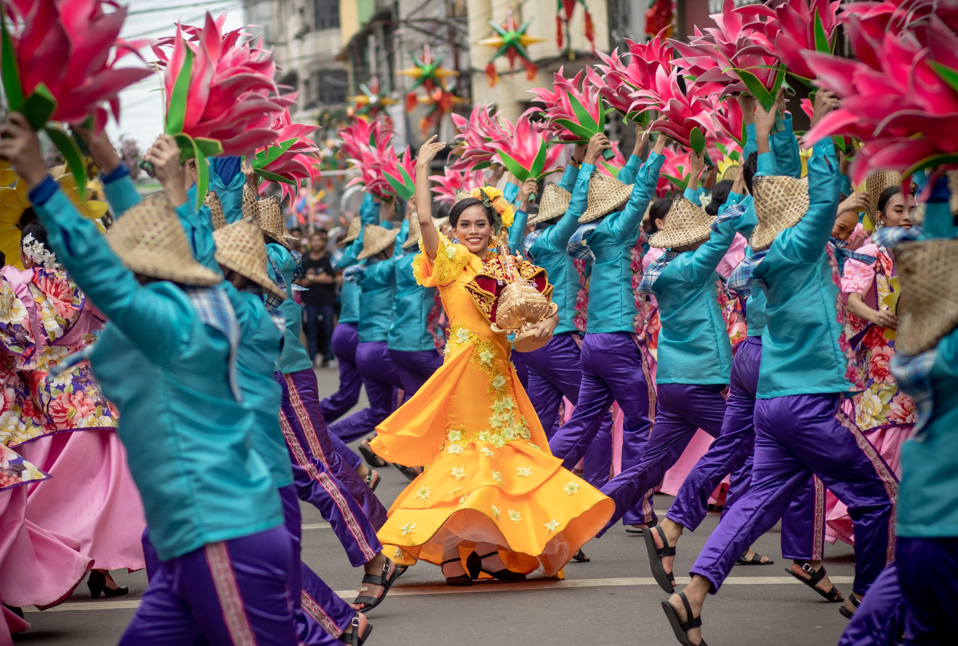 A vibrant festival parade with dancers in colorful costumes and a woman in a yellow dress smiling.