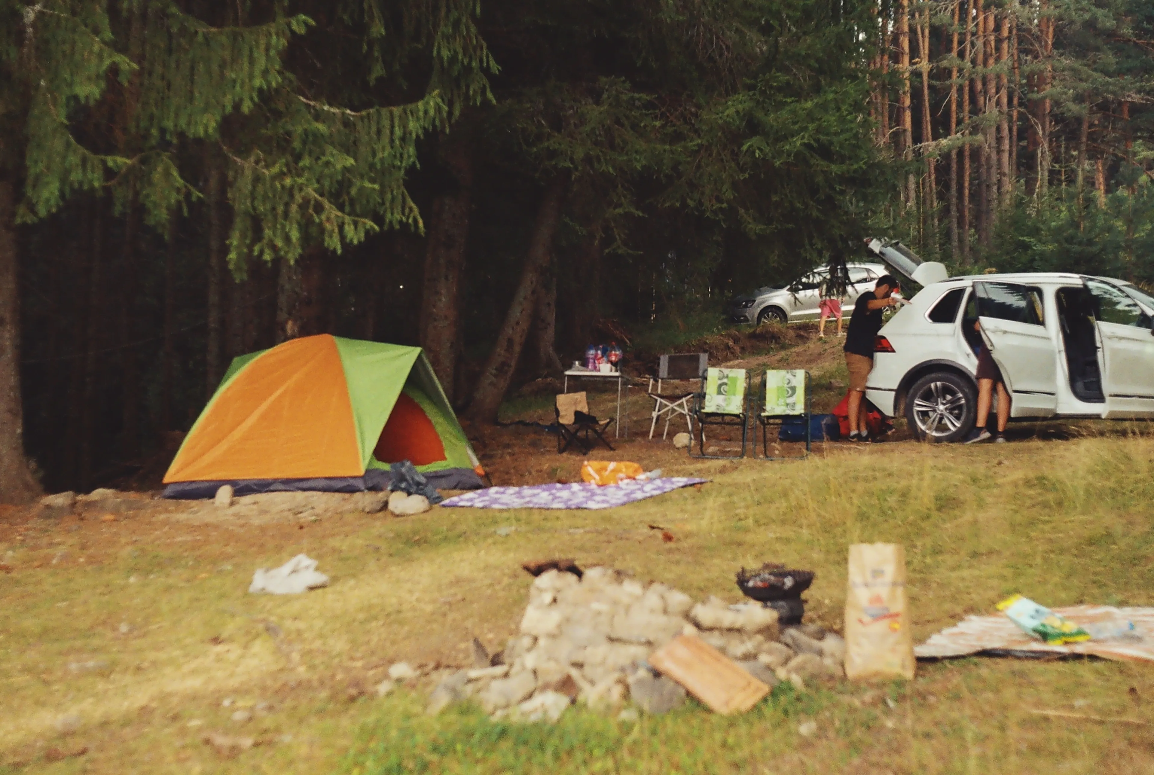 A green and orange tent set up in a forest, with a white car nearby, enjoying a camping trip.