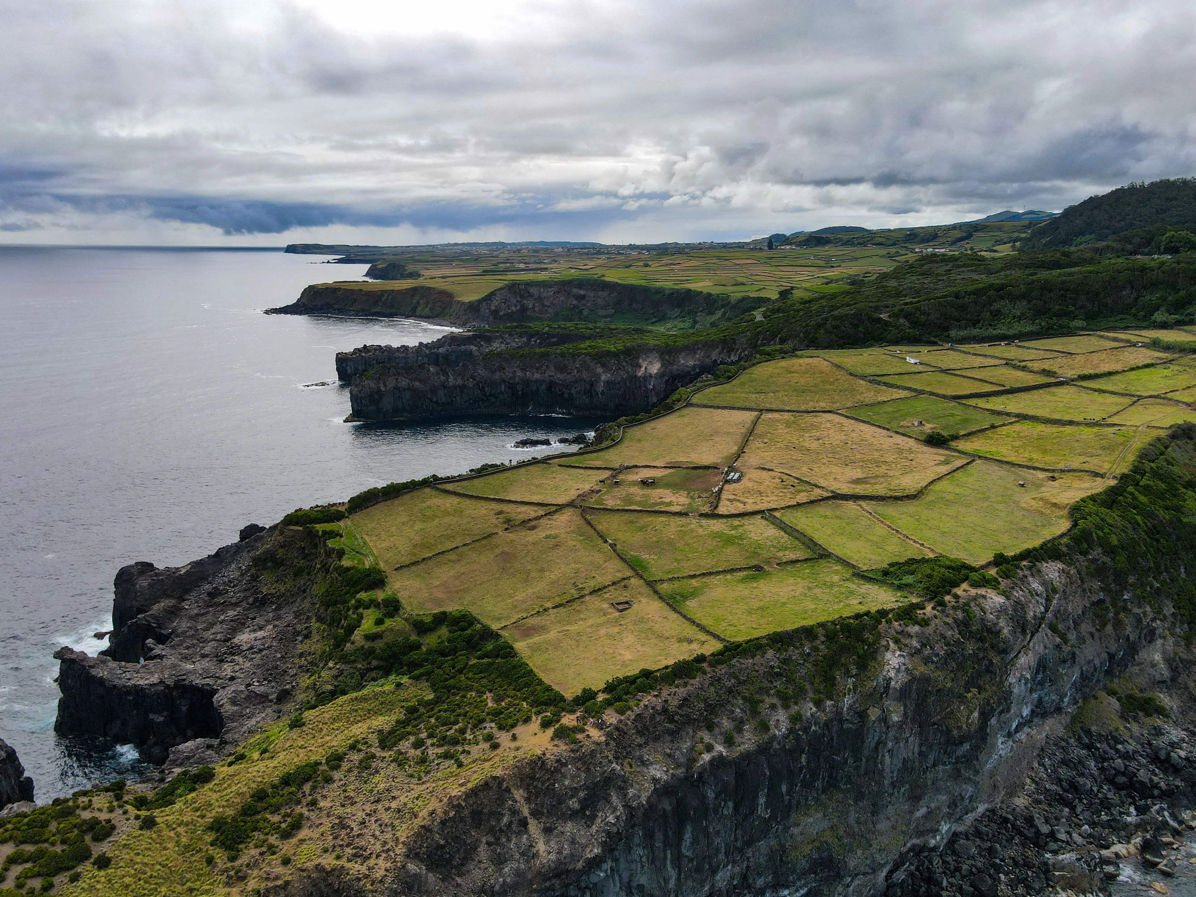 An aerial view of the dramatic coastline of the Azores, with green fields divided by stone walls and ocean cliffs.