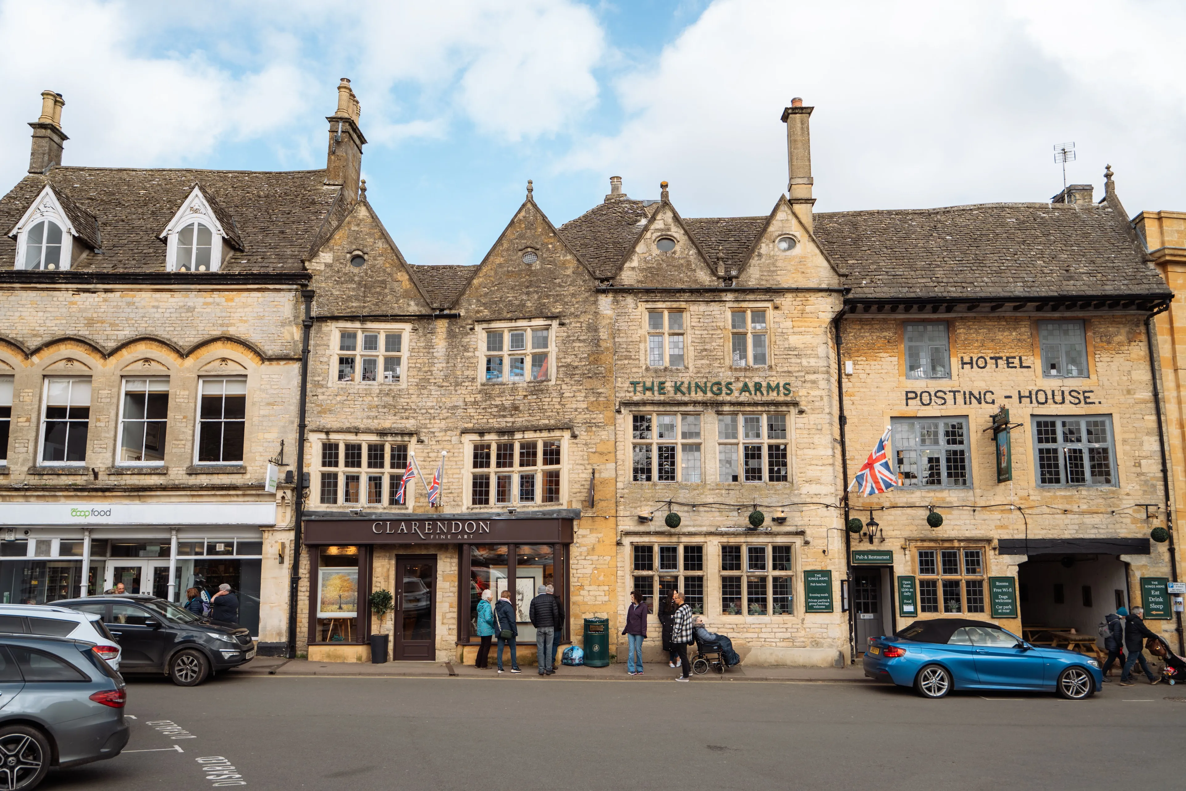 Charming street view of The Cotswolds with historic buildings, a pub, and parked cars on a cloudy day.