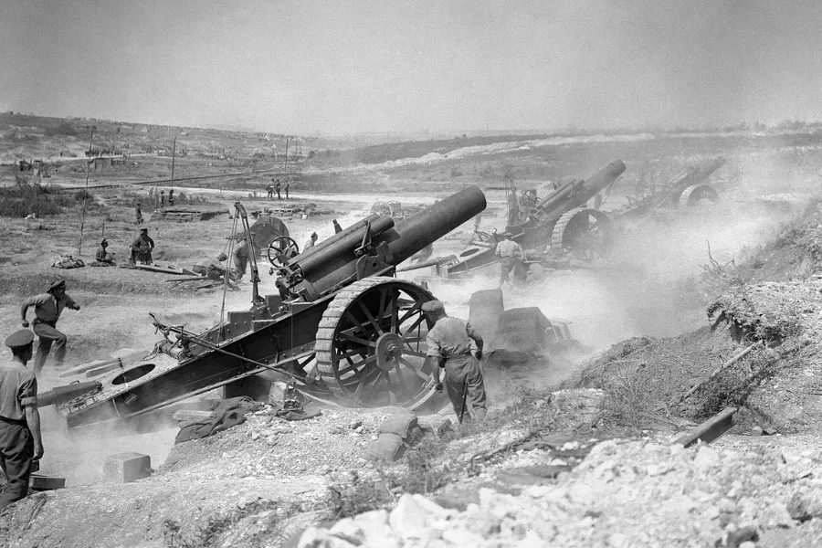 Soldiers operate large artillery cannons in a smoky, open field during the beginning of WWI.