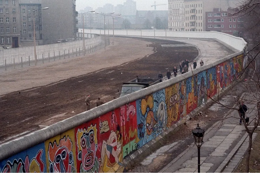 The Berlin Wall is built, stretching across a landscape, adorned with colorful graffiti and people walking nearby.