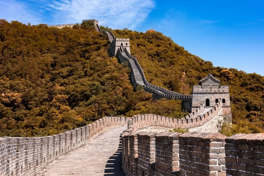 The Great Wall of China winding through mountainous terrain covered with lush trees under a bright blue sky.
