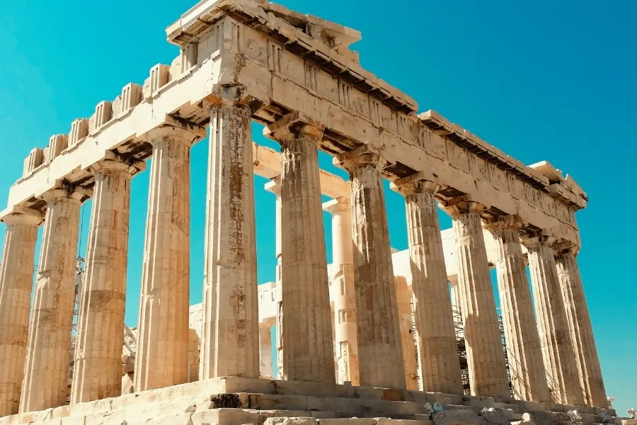 Ancient Parthenon construction with grand columns and intricate details against a bright blue sky.