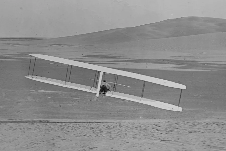 The Wright brothers' first powered flight glider soaring above a sandy landscape, marking a pivotal moment in aviation.