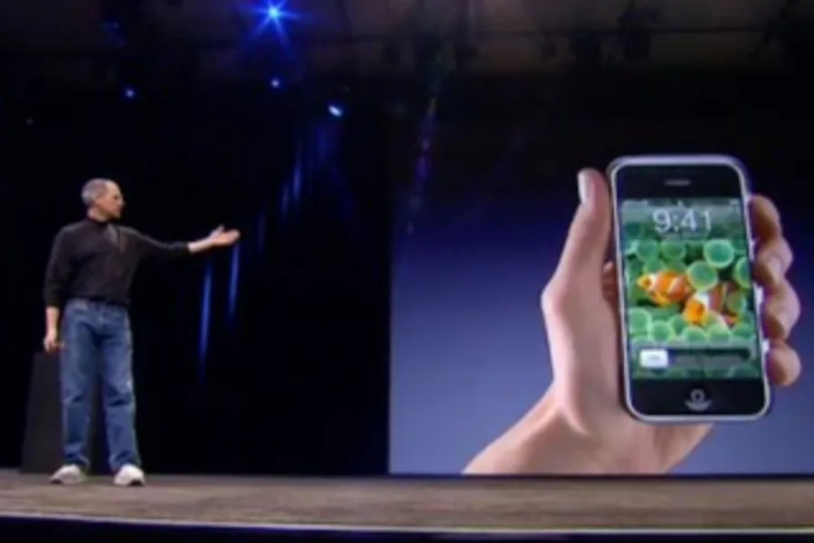 Steve Jobs on stage next to a large screen displaying the Apple iPhone held by a hand, showcasing the release of the Apple iPhone.