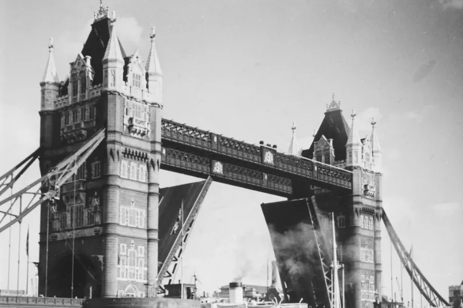 A black and white photo of the Tower Bridge, its bascules raised to allow a ship with smoke emanating from its stack to pass underneath.
