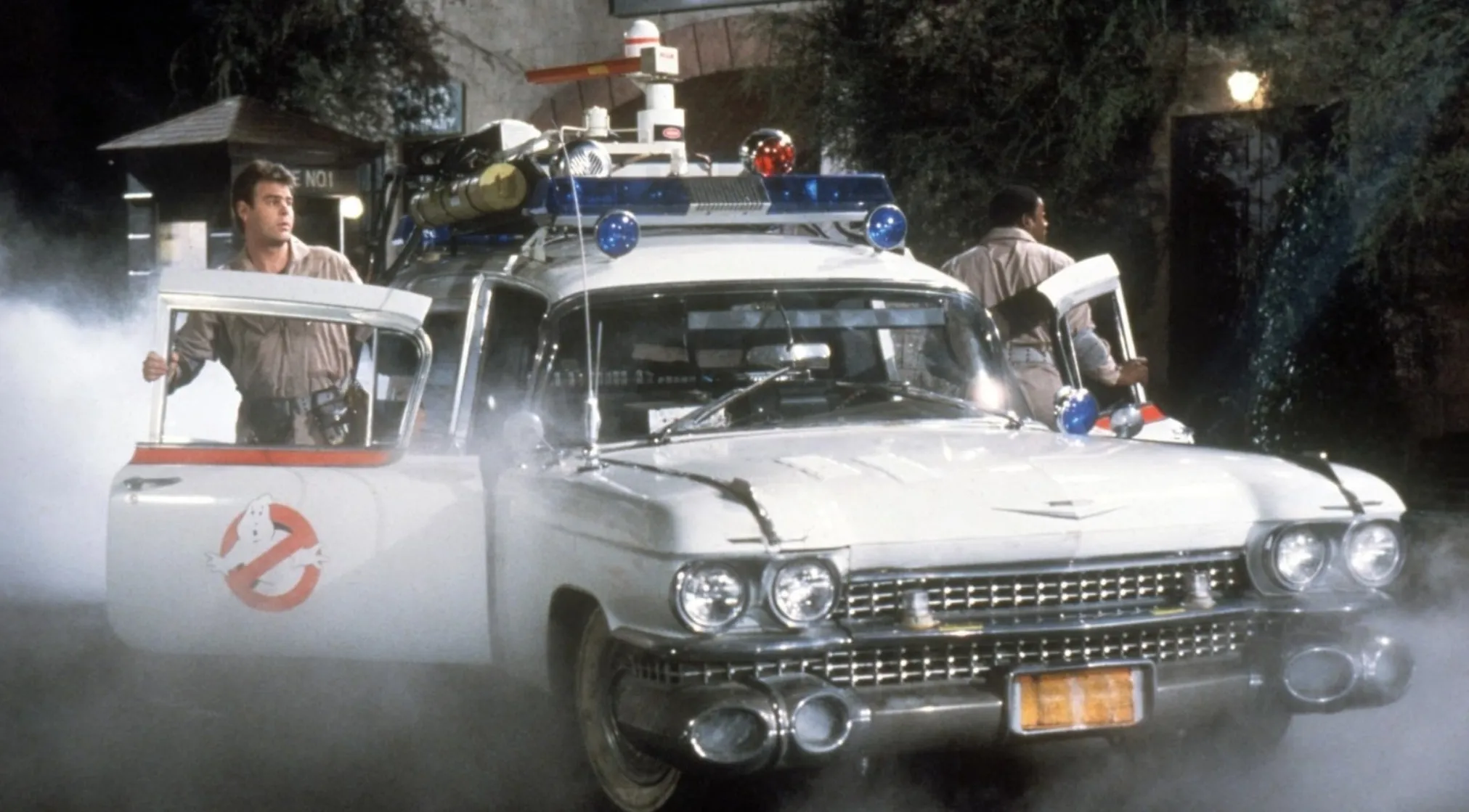 The Ghostbusters iconic vehicle, the Ecto-1, a white Cadillac with flashing lights, emerging from smoke with two crewmen.