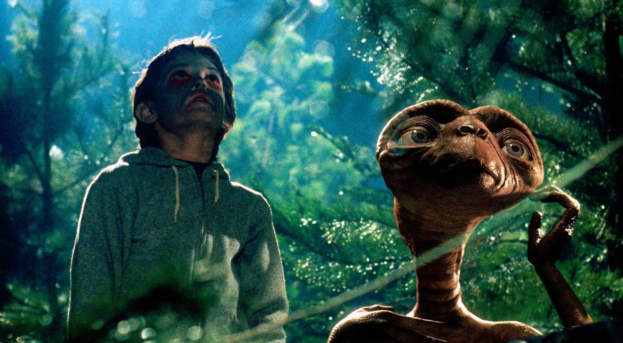 Elliott and E.T. looking up, surrounded by trees. 80s movie trivia fans will remember E.T.'s iconic phrase.