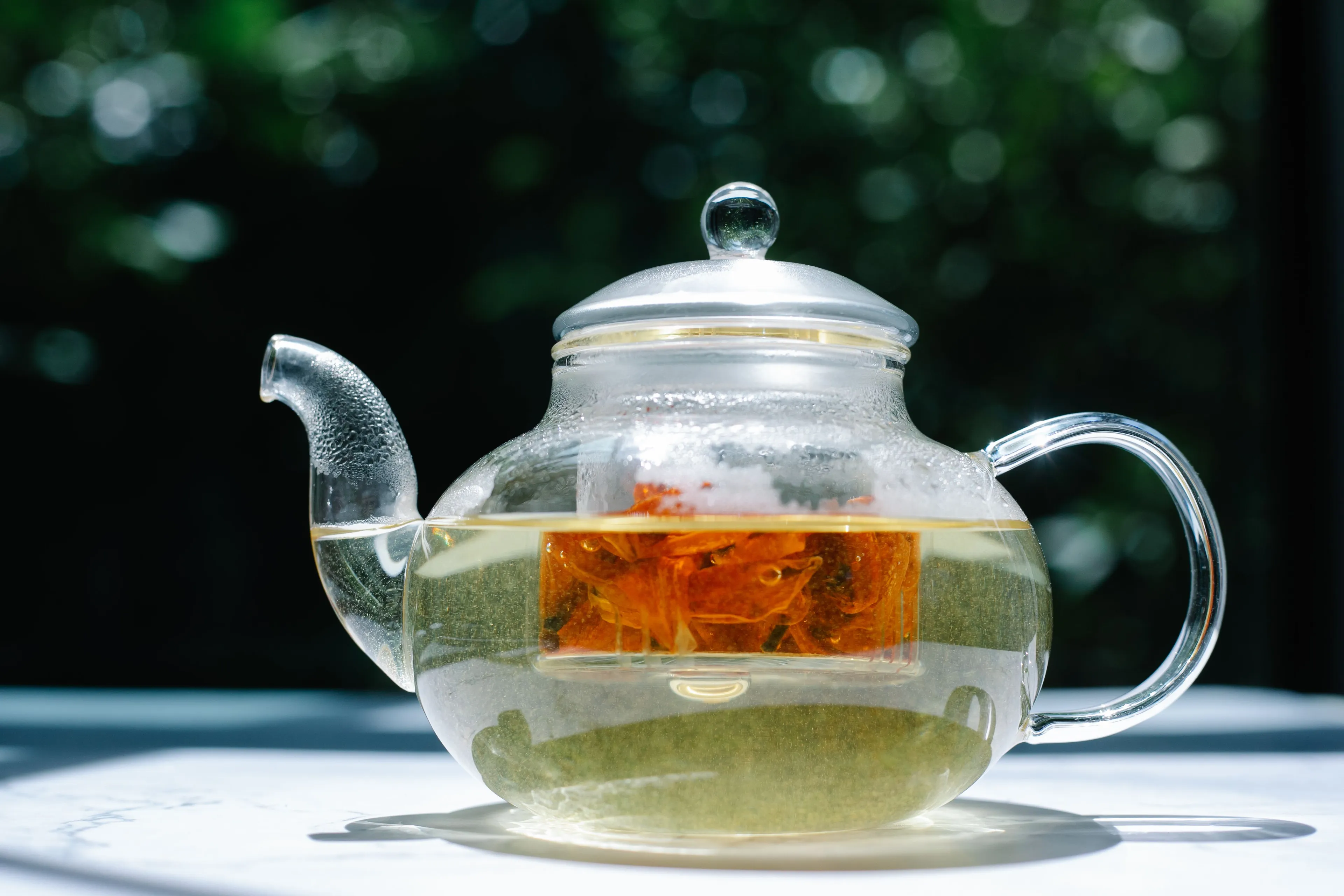A clear glass teapot with herbal tea brewing inside, showing an infuser with a flower, for a drink choice.