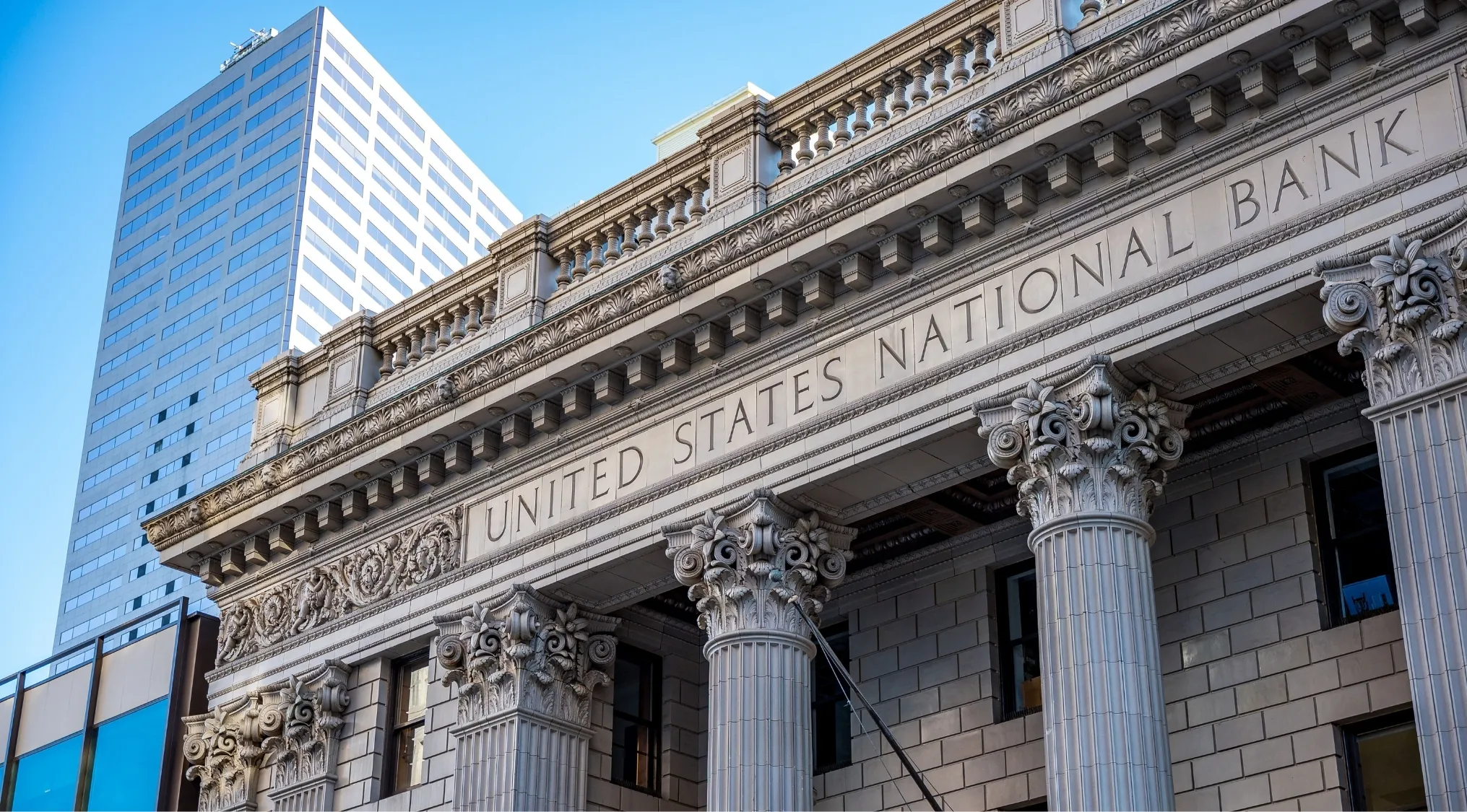 Grand facade of the United States National Bank, with ornate columns and a modern high-rise in the background. Interest arithmetic exam.