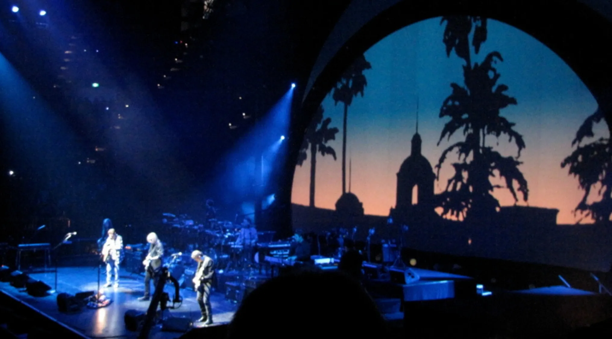 A band performing Hotel California live with palm trees projected on a large screen behind them, part of general music knowledge.