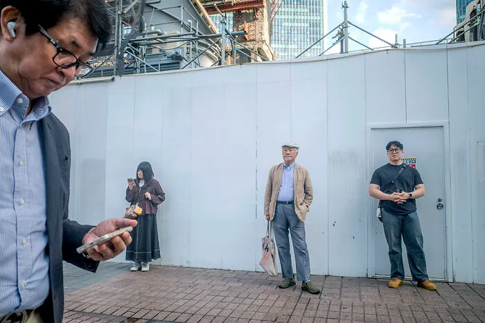 A man engrossed in his phone, framed by a candid street scene in Japan, ideal for street photographers.